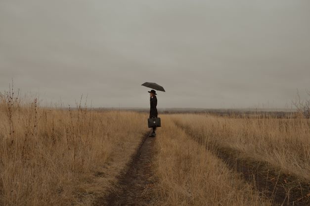 A person in black standing in a yellow golden grass field on a cloudy day holding a black umrella, looking into the distance. Caption: Be a traveler. 
