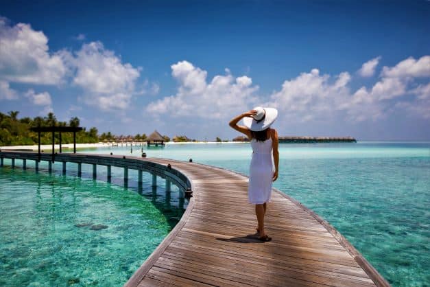A woman walking on an arched wooden pier over crystal clear water on a bright summer day in the Caribbean. She is wearing a white dress, a white hat, and is looking out to sea and the horizons blue skies. 
