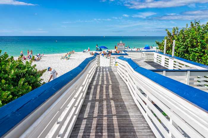 Welcome to Clam Pass Beach; a wooden pathway with white and blue railings leading to the white sands with many sun-lovers, in front of the greenish-blue ocean and the blue skies