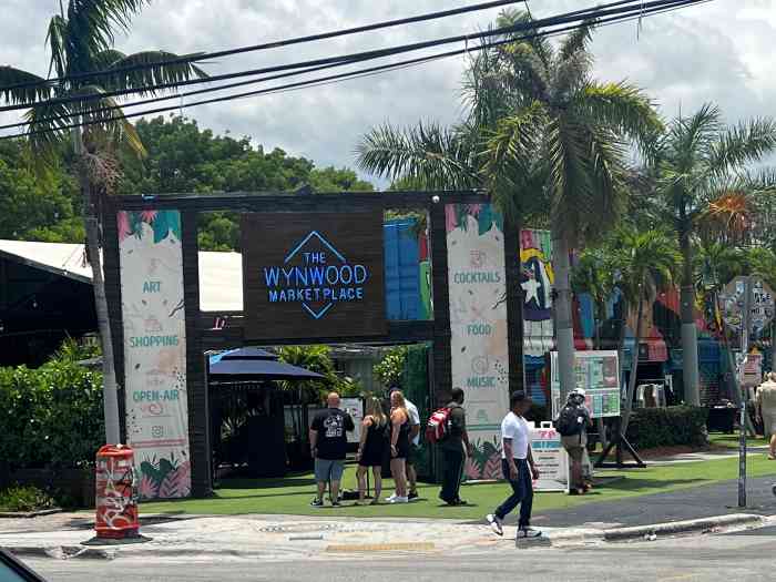 The entrance gate to Wynwood Marketplace on a sunny summer day with people on tehir way in between the wide doors on the green grass. 