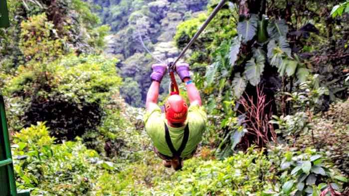 A man on his way zip-lining down the wire in the middle of lush green forest in Costa Rica, wearing a helmet, thick gloves and holding on to the handles of the zip line. 