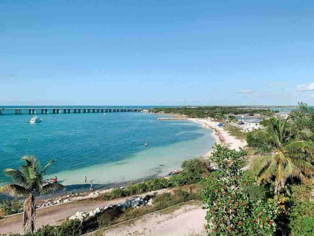 The sandy bay at Bahia Honda State Park on Marathon Key, Florida, surrounded by palm trees, and you can see the Seven Mile Bridge in the distance under the blue sky. 
