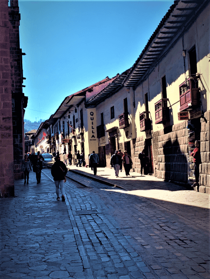 Everything is stone in Cusco. This street has stone for pavement, and the beautiful buildings around it are light brown and yellow stone. The sky above is blue. 