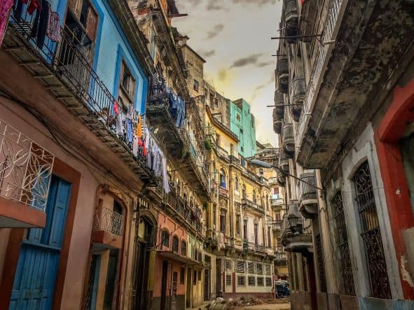 A narrow street in Central Havana, with intricate architecture on run-down houses that are painted in a variety of pastel colors. The balconies are decorative and are filled with laundry hanging to dry, the doors are in all colors and shapes, and the same with the windows that are often covered with artfully crafted iron rails. 
