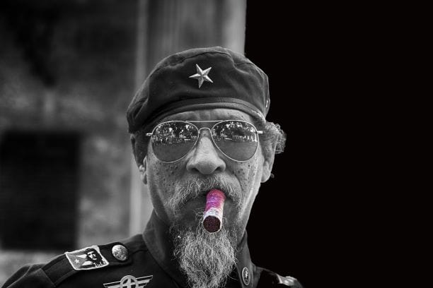 Black and white photo of a man with a huge Cuban cigar, wearing a military uniform, with a beret and pilot sunglasses. 