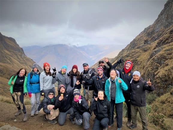 The hiking group together on the top of Dead Womans Pass on the Inca Trail, all smiling and happy to have made it this far! There are green hills on both sides, and infinite views of the mountains in the background. 