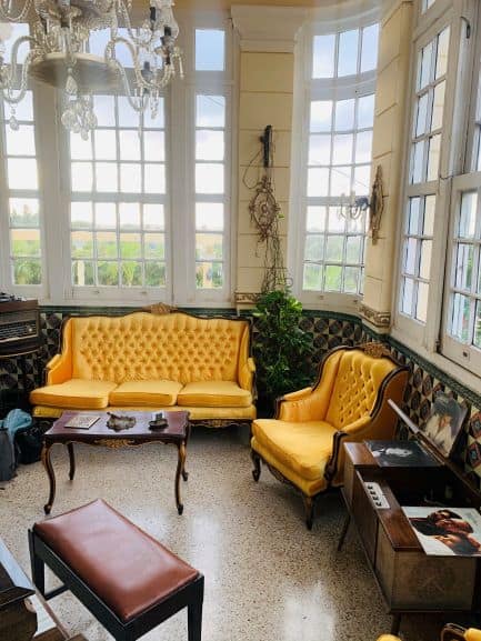 A living room in Villa Teresa in what is almost a winter garden with large windows towards the garden. The rococco sofa is bright yellow, and surrounded by chandeliers, green plants, and art deco furniture. 