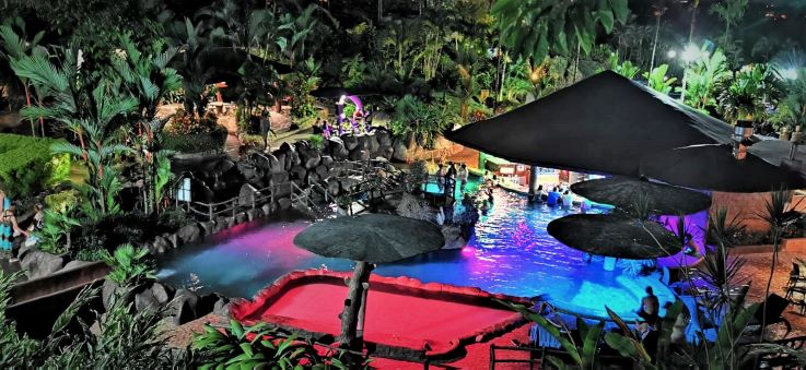 Hot springs in Costa Rica at night, the pool is lit with blue and purple lights, and people are enjoying themselves in the swim-up bar surrounded by stunning greenery