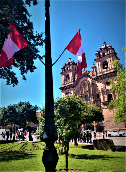 Plaza de Armas in Cusco and the church, the park is decorated with Peruvian flags, the church is in warm beige stone, with elaborate decorations, and the sky is blue behind it all. 