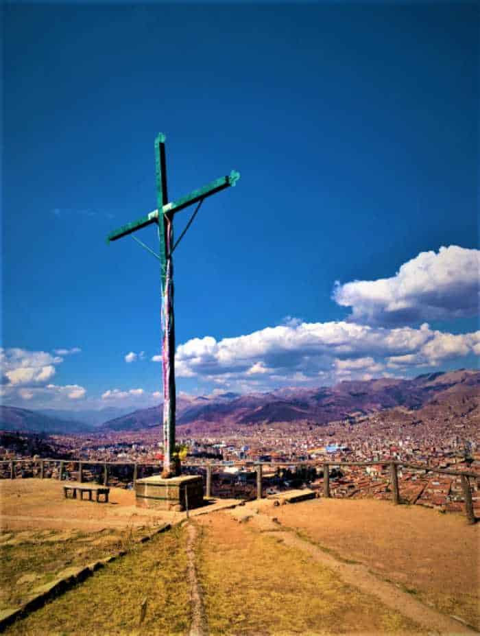 The tall cross of Christ standing on a dry grassy plain high above Cusco with the city roofs down below and the mountains in the distance on a sunny day