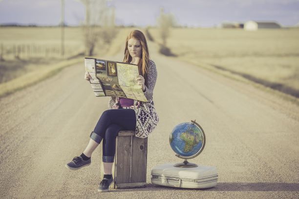 Woman sitting on her suitcase on a dusty road, reading a map, with the world globus beside her. 