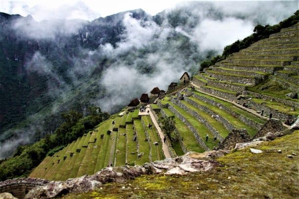 Incredible green terrace structures sitting on a steep hill along the Inca Trail. You see floating dots of cloud in the background against the dark green hills. 