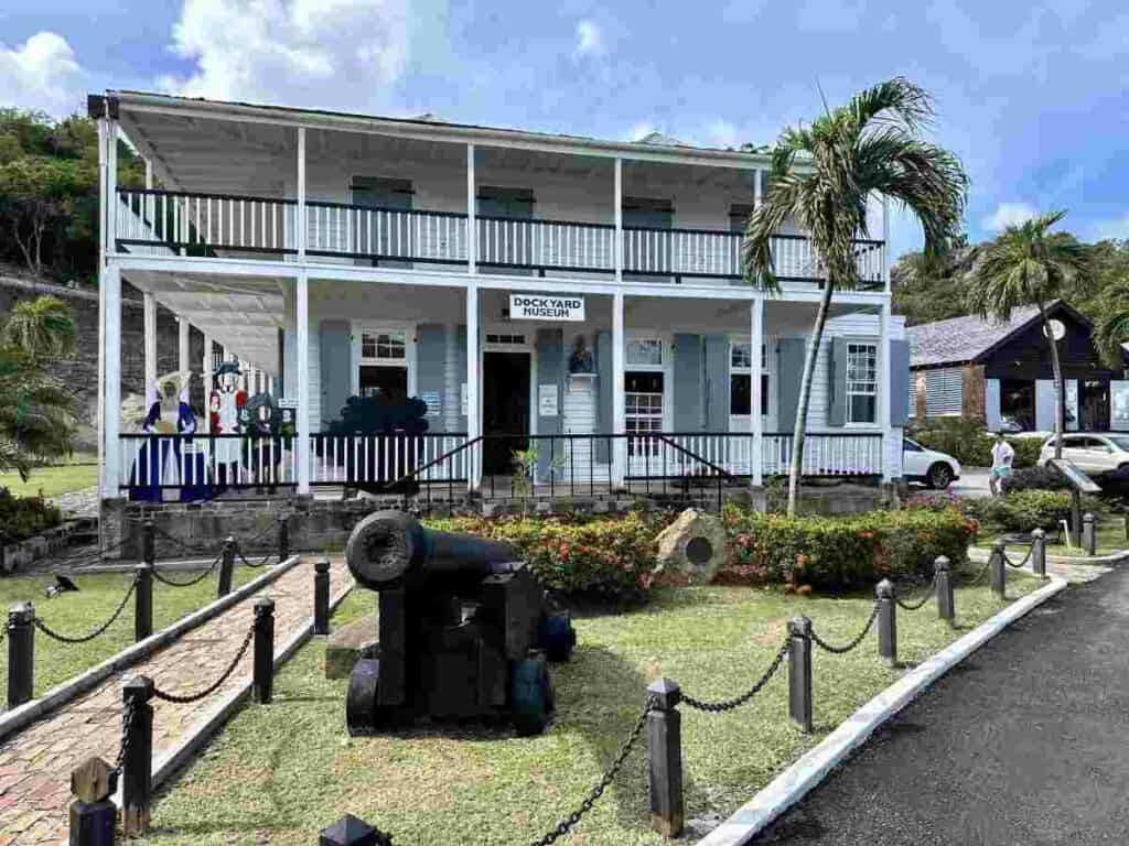 Historic white and grey buildings at Nelsons Dockyard with a green lawn in front, bushes and flowers and even a small black old cannon on a sunny summer day