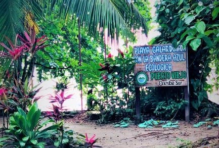 The entrance to a beach in Costa Rica on a path through lush green plants, in a place called Puerto Viejo like the sign says in the photo, along the Caribbean coast