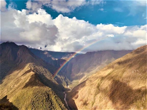 Beaufiful views of endless valleys, with a rainbow straight across the infinite distance. The sky is blue, the hills are green and yellow, and there are a few white clouds. 