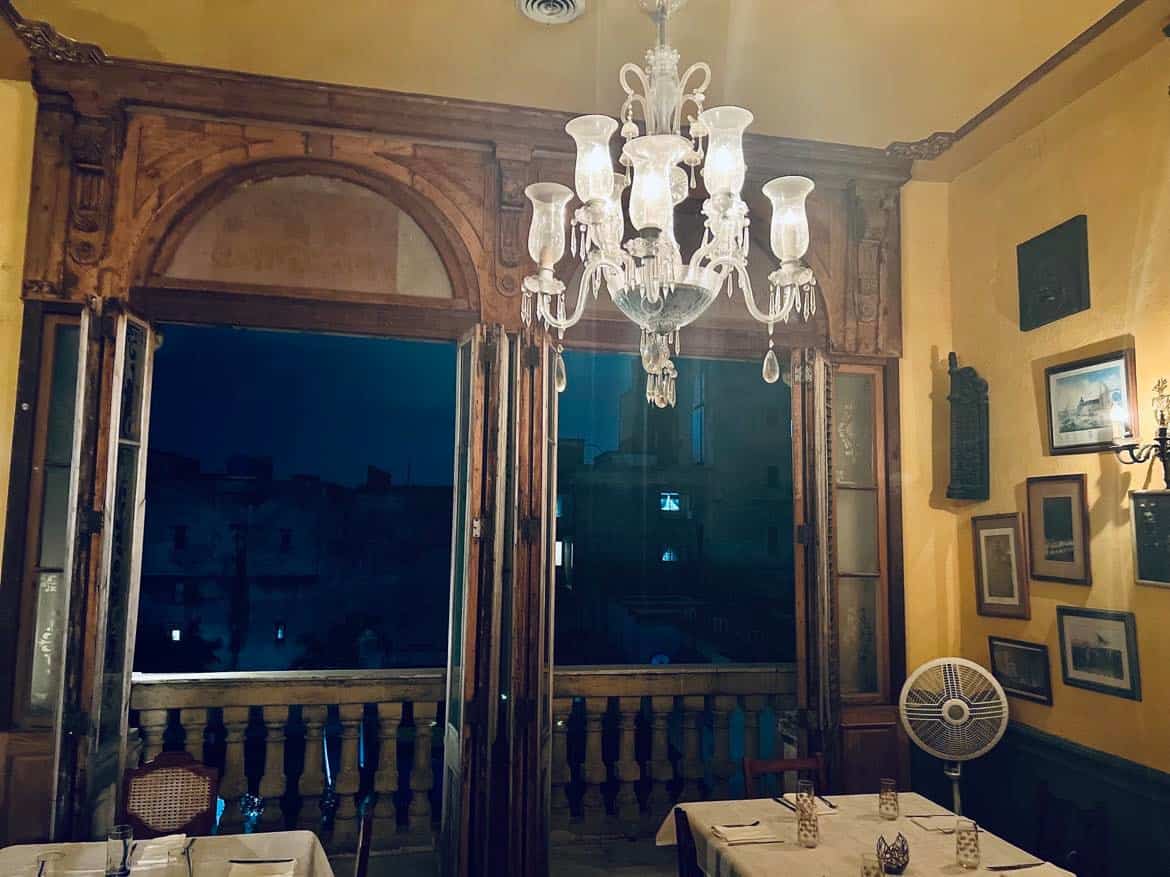 Beautiful dining area in La Guarida, Central Havana, with a large glass chandelier, yellow painted walls, wide open windows with wooden palistrades behind the white clothed tables. 