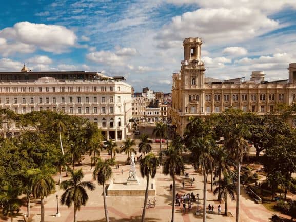 View of the Parque Central in Havana on a summer day, with lots of palm trees and people, surrounded by beautiful classic colonial elegant buildings. 