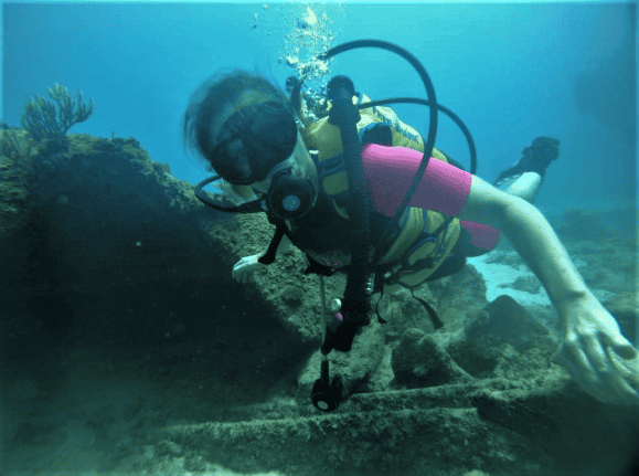 Close up photo of me scuba diving in Havana, trying to smile behind the mask. Wearing a pink wet suit, holding on to the remnants of an old wreck, crystal clear water all around me. 