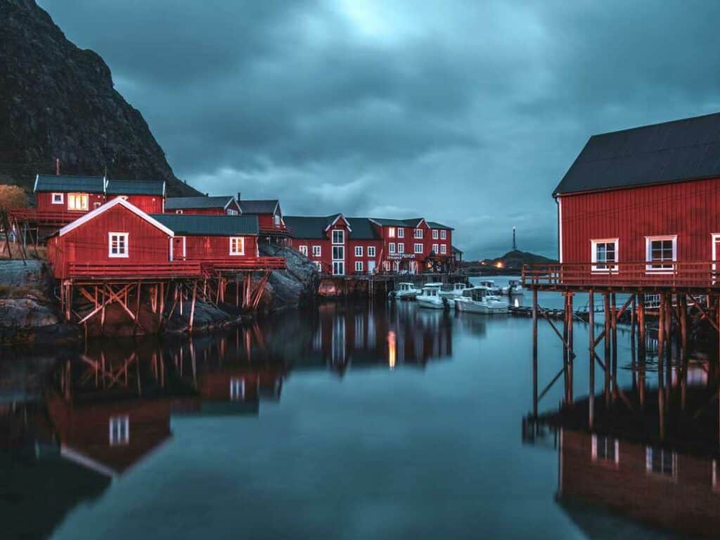 Clouded evening in Å in Lofoten, with calm dark waters under the bright red wooden cottages sitting partly over the water beside the vast mountains