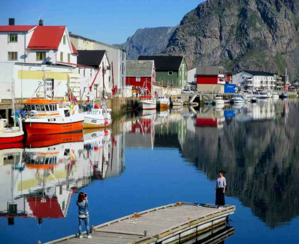 Henningsvær on a sunny day, with blank calm water surrounded by white and colorful wooden houses under the blue sky and beside the steep mountains