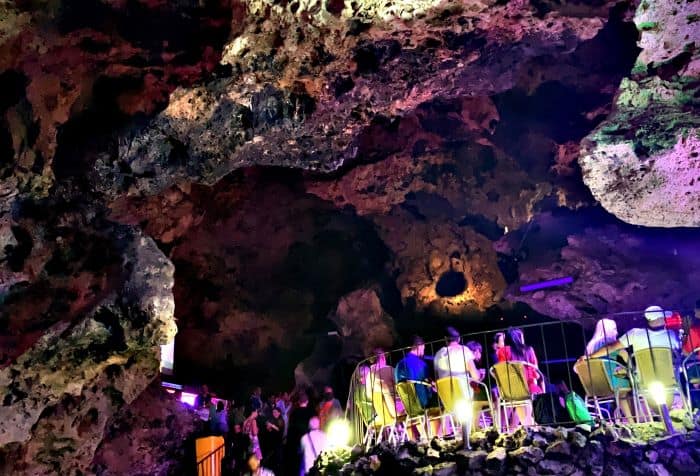 Seating area in La Cueva night club, on an elevated sorky place under the rugged cave walls and ceilings, people are sitting on bright yellow chairs amidst the colorful lighting