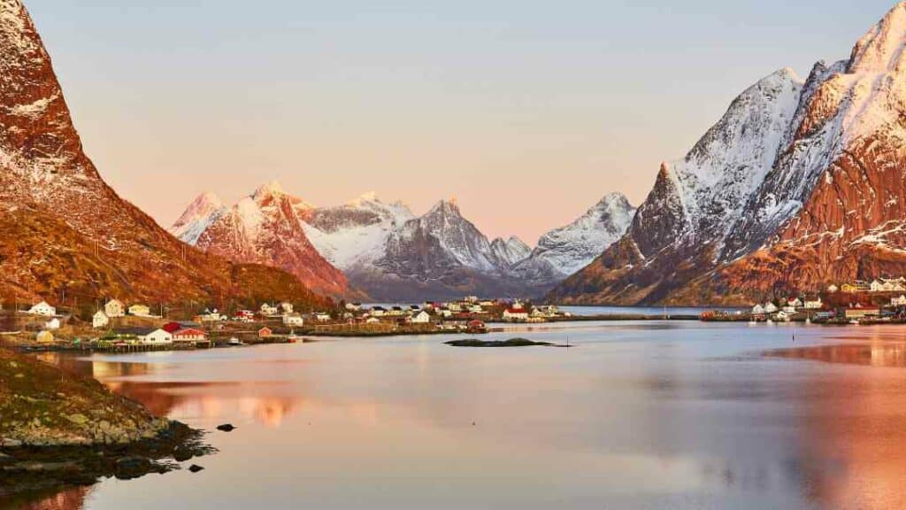 Lofoten Norway, a fjord bathed in warm sunlight amidst steep mountains around a quaint village with the snow capped mountains visible in the distance.