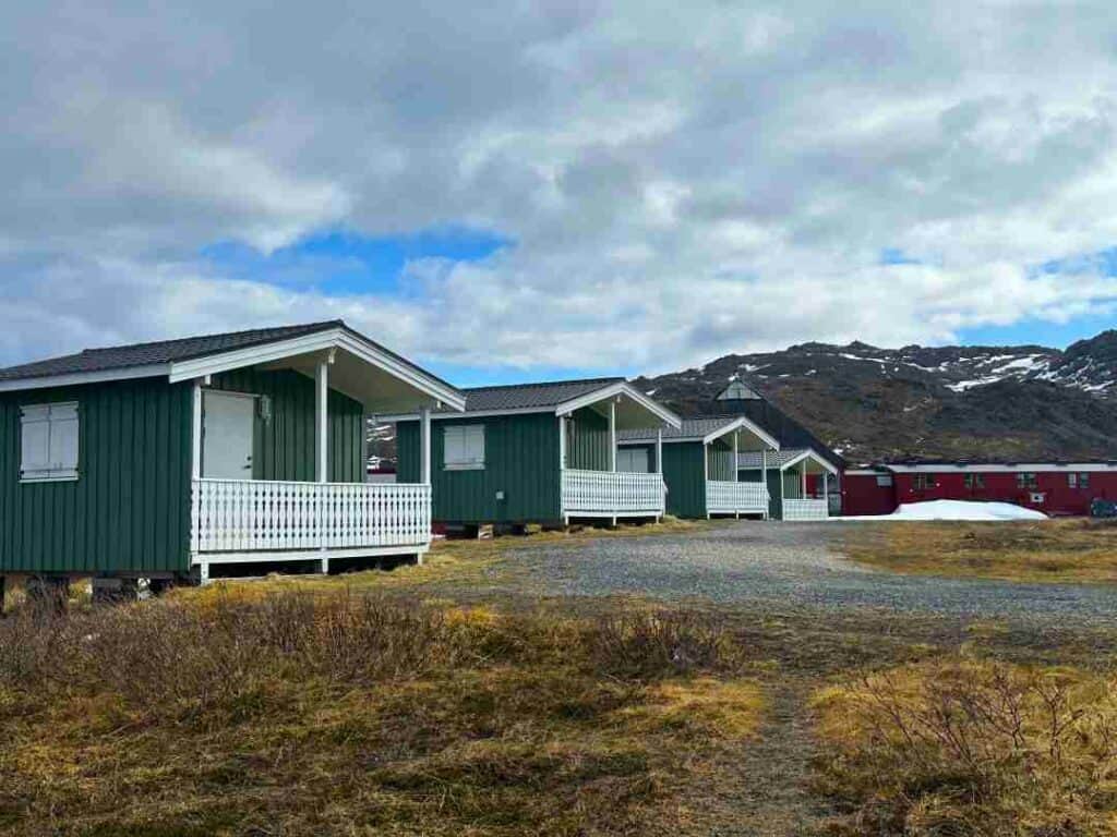 Small green camping cabins outside Honningsvåg in May on a partly cloudy day when there still is a bit of snow, and the grass is yellow, with the wavy mountains in the distance