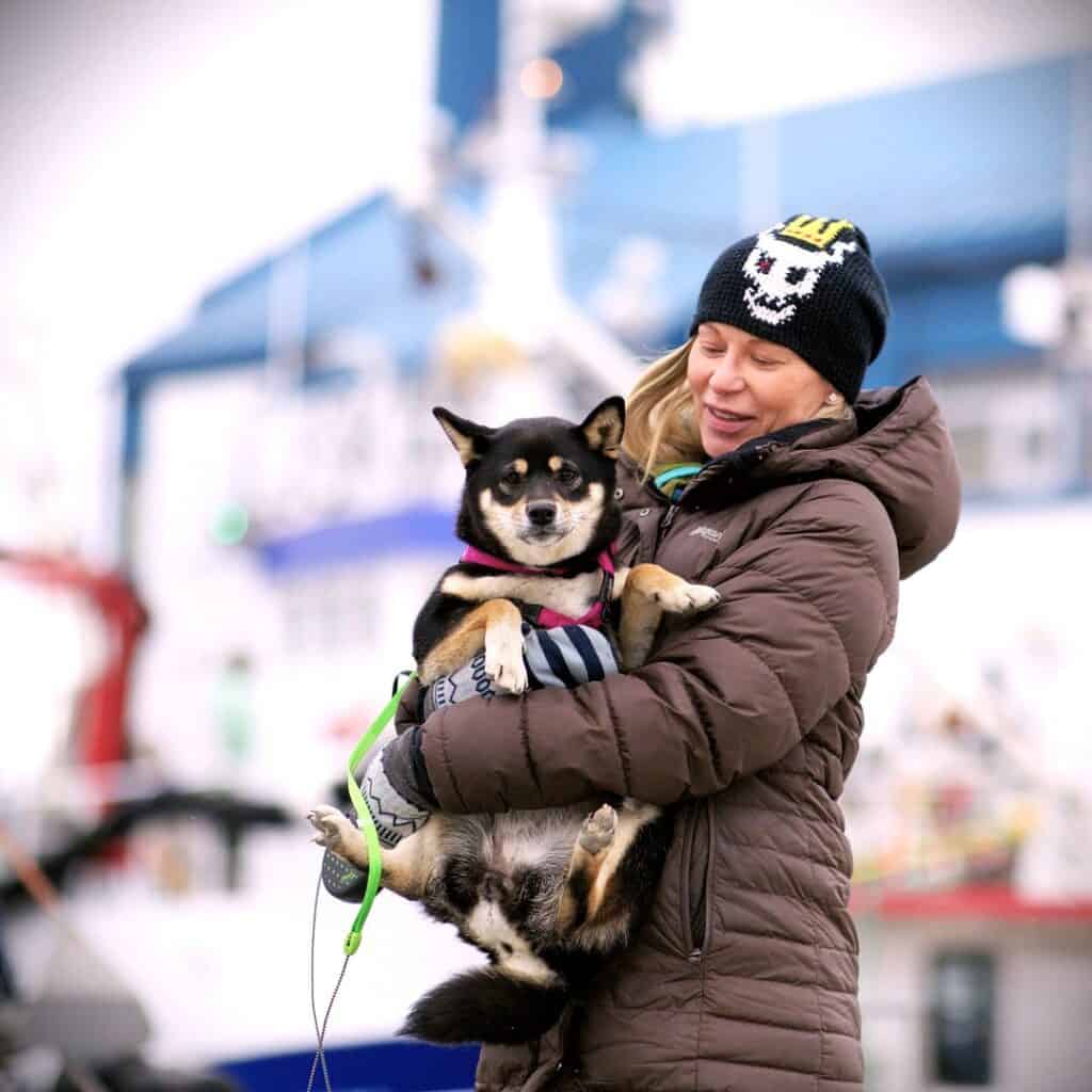Me wearing a long insulated jacket and hat in May in Honningsvåg, holding a cute dog while visiting the docks in Honningsvåg on a cloudy but bright day. 