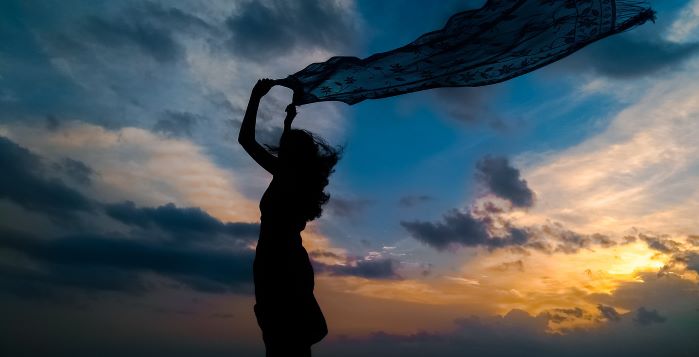 Lady in the sunset holding a scarf over her head that is flowing in the wind under a blue sky with golden rays from the setting sun