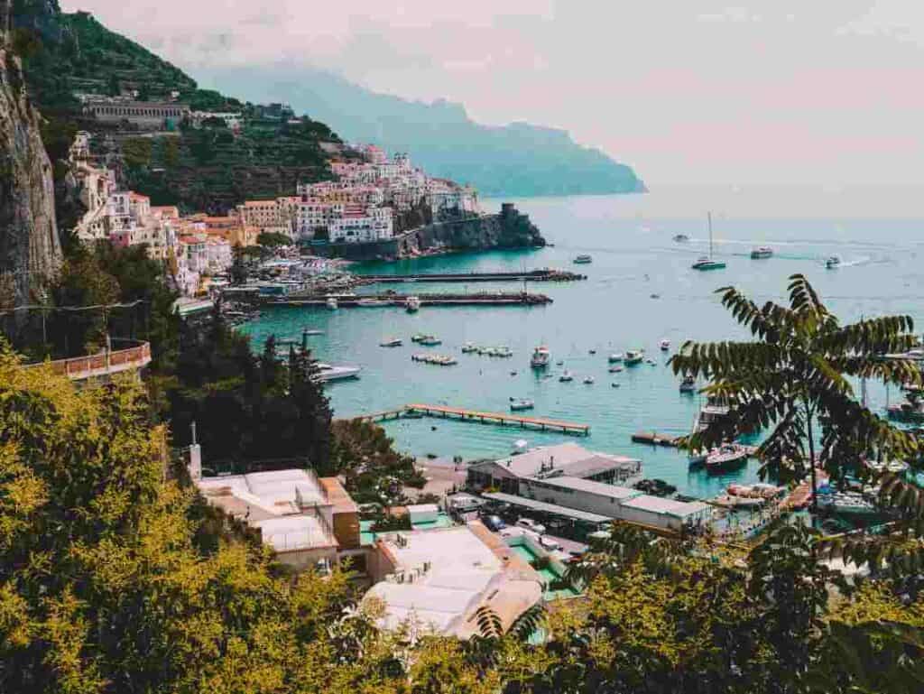 The incredible Amalfi coast in Italy with houses clinging to the steep mountain side above the water, the narrow streets, the harbor with green water and boats, and hills in the background