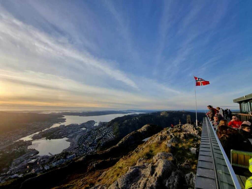 People standing on a viewpoint terrace looking down at a town far below, and the infinite view of the fjords and the sea under a blue sky