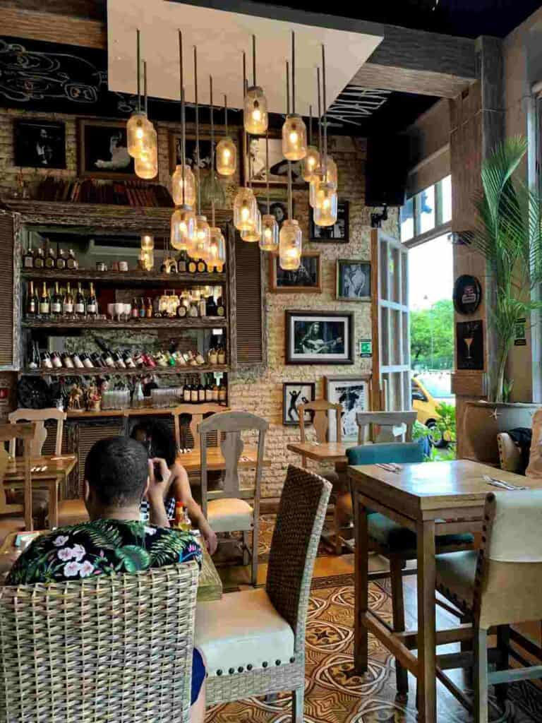 Is Cartagena Colombia Worth Visiting? A super charming cafe in the center of Cartagena, with brick walls, wooden details, green flowers, warm lighting, photos and decorations beside the wine shelf and charming furniture. 