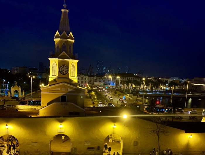 The view from the famous yellow clock tower at night in Cartagena, with warm lighting around the tower, and the city lights in the distance behind it