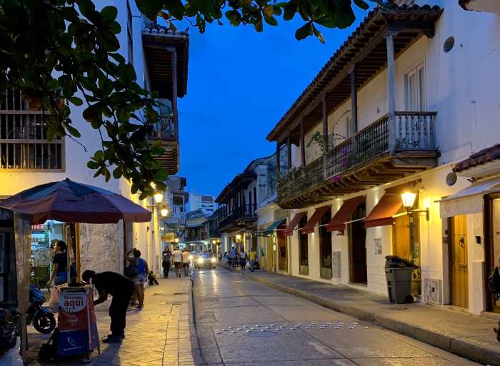 A charming old city street in Cartagena at night with a deep blue sky above the colonial buildings, people strolling and artsy details on teh doors, windows and balconies of the traditional buildings. 