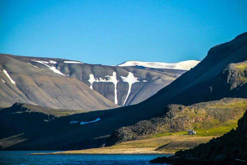 Svalbard in summer, with the characteristic mountains, claciers still on the mountain tops, and green vegetation in the summer sun in the lowland behind the brownish grey mountain sides under the blue sky