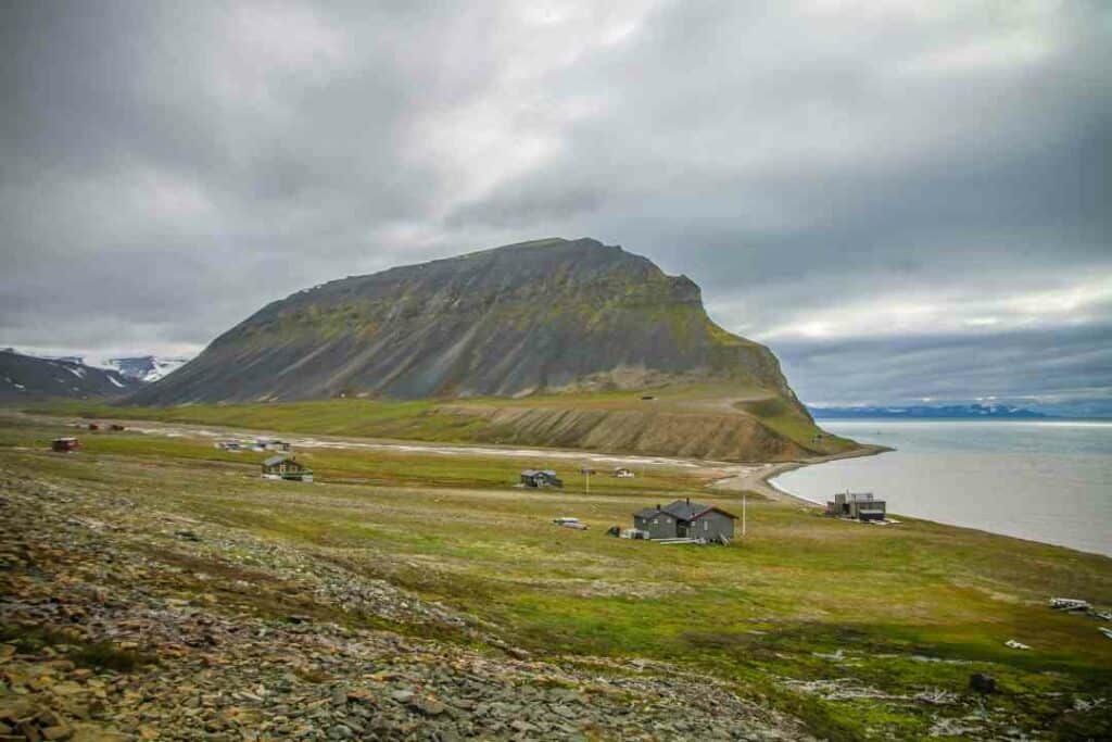 Desolated area called Jan Mayen in Svalbard in September, with whitish grey cloud cover, still green fields with some yellow, and scattered houses along the sandy shores and the mountains