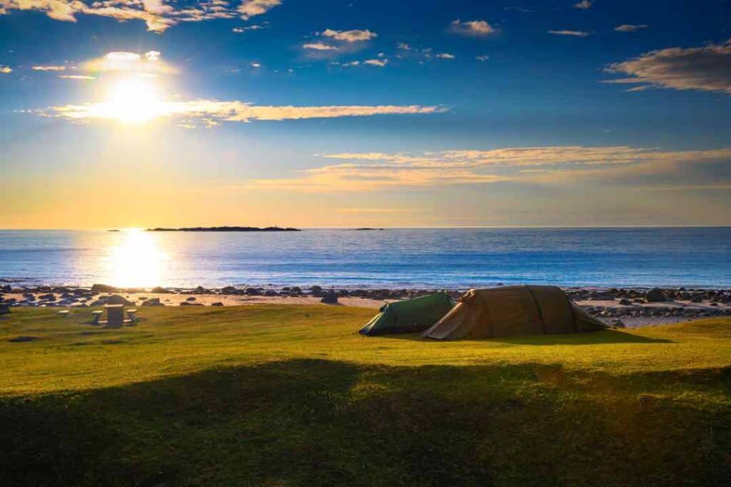 A beach at sunset, with green grassy plains in front of the sandy beach, and two tents on the grass in the warm sunlight from the setting sun over the sea and the horizon