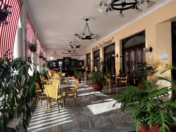 An elegant outdoor patio with green plants, and charming wooden seating areas next to Parque Cespedes square in Santiago de Cuba on a sunny day, the sun hitting the marble floors between the furniture