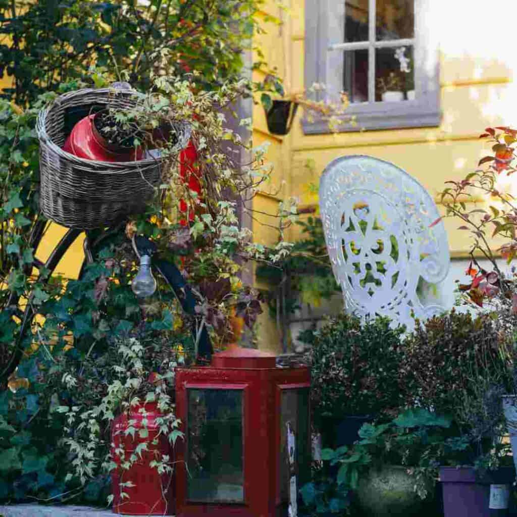 An outroor seating area in Bergen in the summer with lots of flowers and greenery, a white ornate chair, a bike covered in flowers and lights in front of a yellow wooden house. 