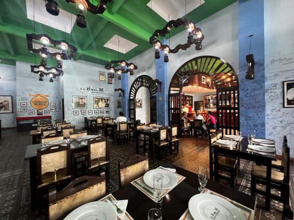 The Bodeguita del Medio interior in Santiago de Cuba, with green ceiling, blue and light blue walls, lots of memorobilia and art, brown wooden solid tables and chairs ready with white plates and wine glasses