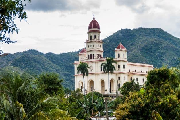 The beautiful white church called El Cobre Sanctuary outside Santiago de Cuba, with towers with decorative red domed roofs, towards a backdrop of the lush green hills