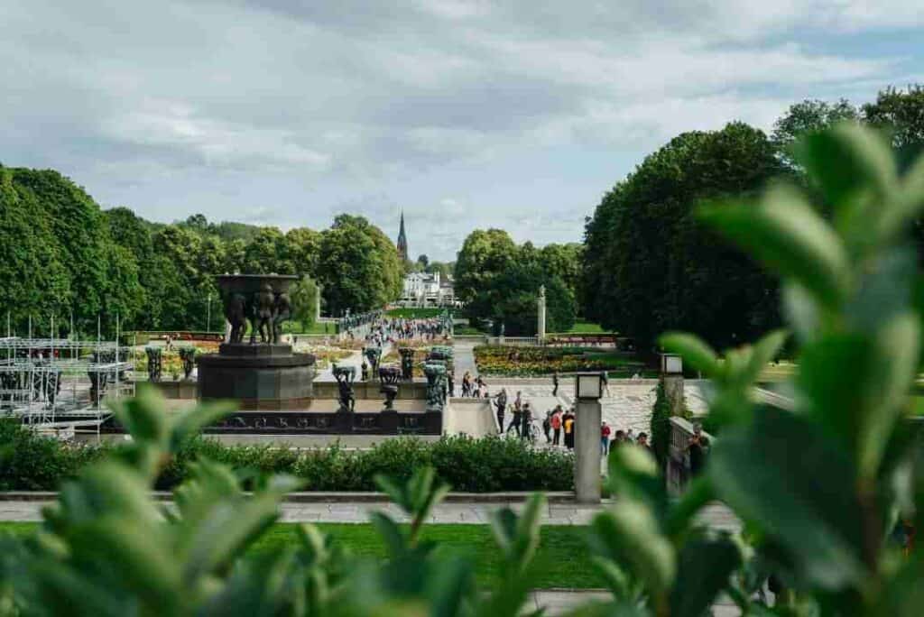 The Frogner Park, also called Vigelandsparken, on a summer day with lots of people, lush green trees, flowers, and stunning pieces of art between the greenery