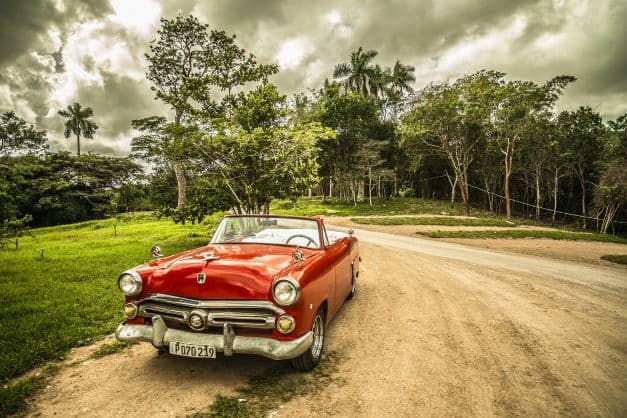 A shiny and bright red classic American convertible car in Cuba, standing on a dirt road in the middle of a green forest