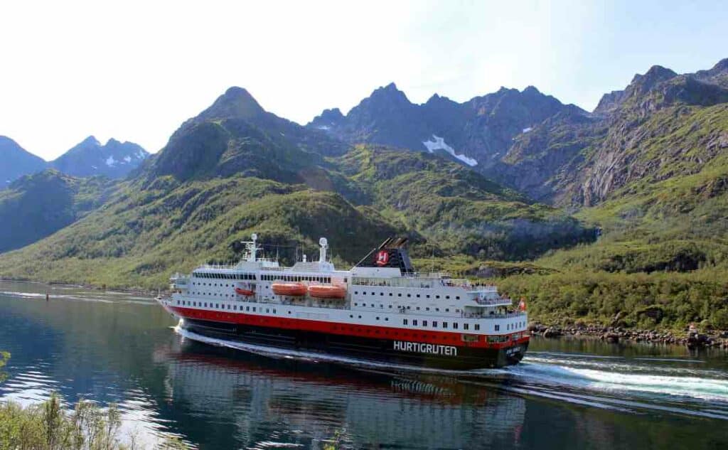 The red, white and black Hurtigruten coastal ferry starts and ends it journey along the Norwegian coast in Bergen. Here it is in a fjord below green mountain sides where there still are patches of snow on the peaks on a sunny day