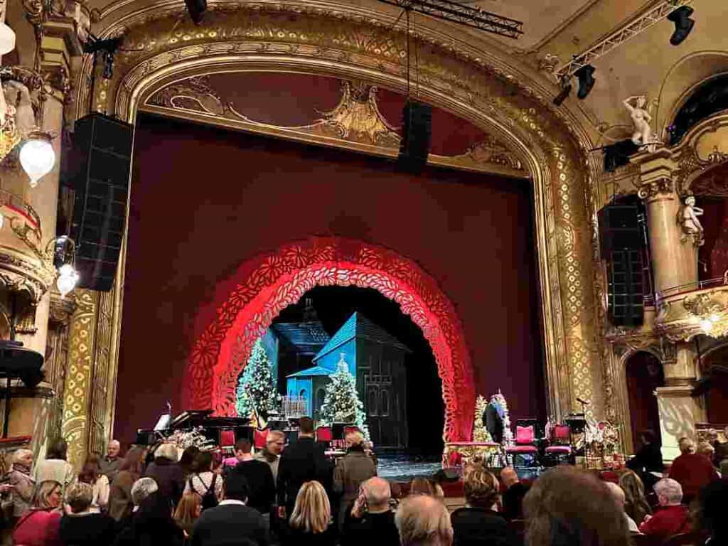 The National Theater Stage in Oslo, with golden decorations, columns, high ceilings, elaborate art, and lots of people waiting for the performance to begin on stage that is draped in deep red