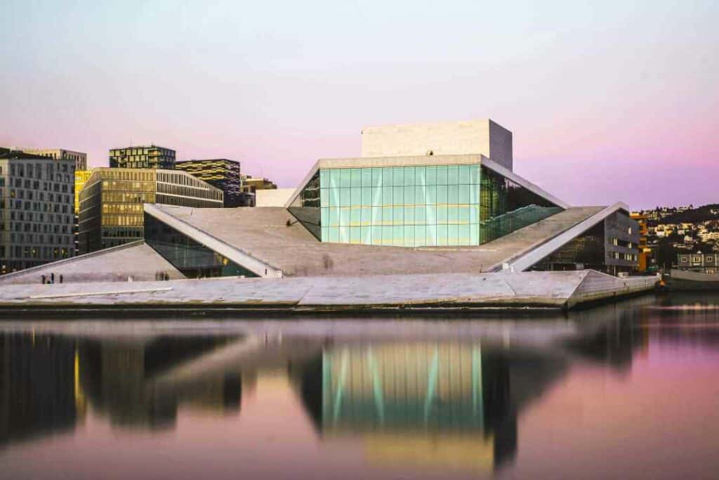 Oslo Opera House from the sea with the green glassy facade at sunset, the sky light bluish pink and the water dark with pink reflections