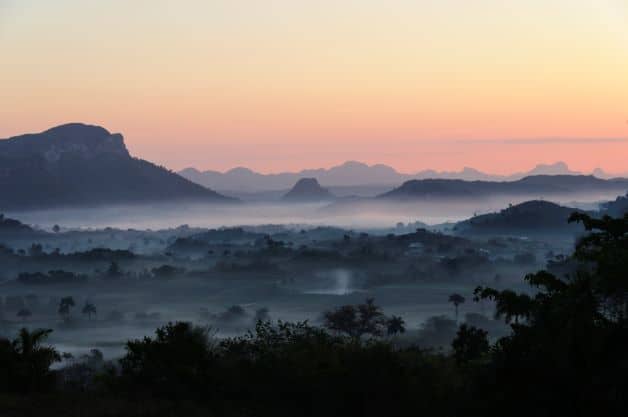 Stunning misty sunrise over Vinales Valley with a glowing pink light in the horizon, silvery mist floating around the valley floor around the trees, and mogotes seen from above