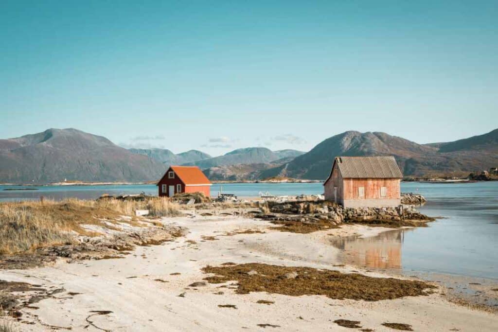 A white sandy beach with sea weed here and there, two red wooden cottages and barren mountains in the background against a pale blue sky