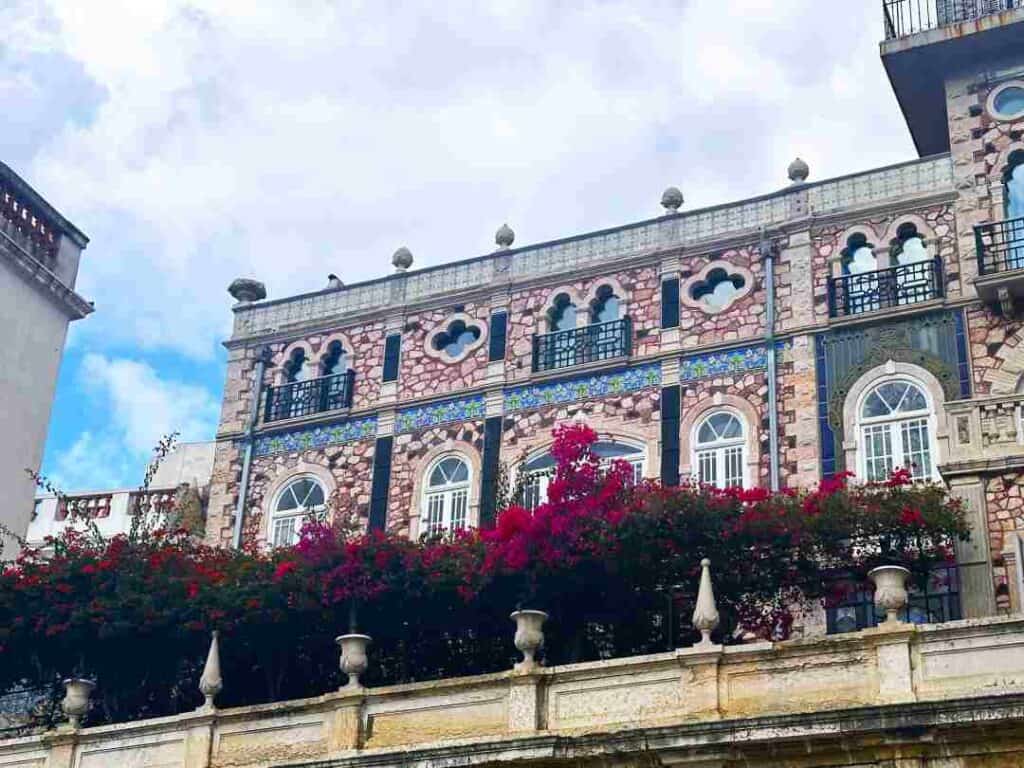 An old stone and tile building with intricate art details on the walls surrounded by greenery and bright pink flowers