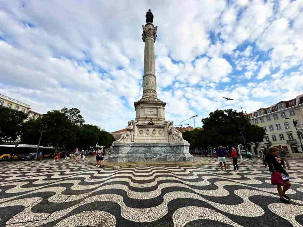 A statue standing high on a white column in the middle of a square with trees and people